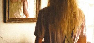 affiche du film La dernière nuit de Lise Broholm de Tea Lindeburg : une jeune femme aux longs cheveux blonds, de dos face à un miroir au-dessus d'un lavabo. Sur le mur à côté, une croix chrétienne.